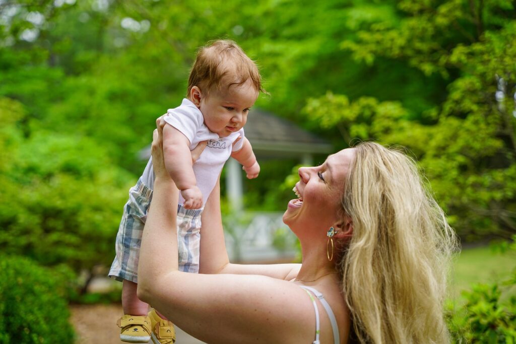 An adult lifting a smiling baby outdoors in a bright, green garden setting.