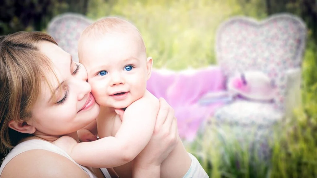 An adult holding a smiling baby outdoors in a soft, natural setting.
