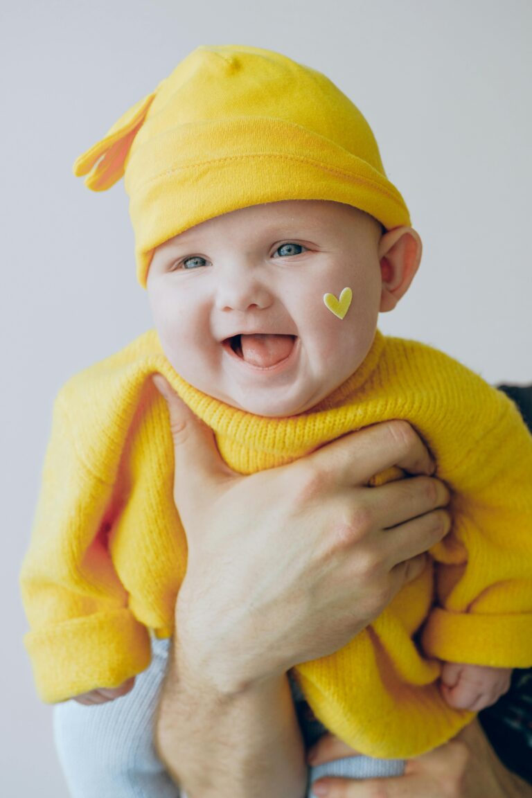Laughing baby in a yellow hat and sweater held up by an adult