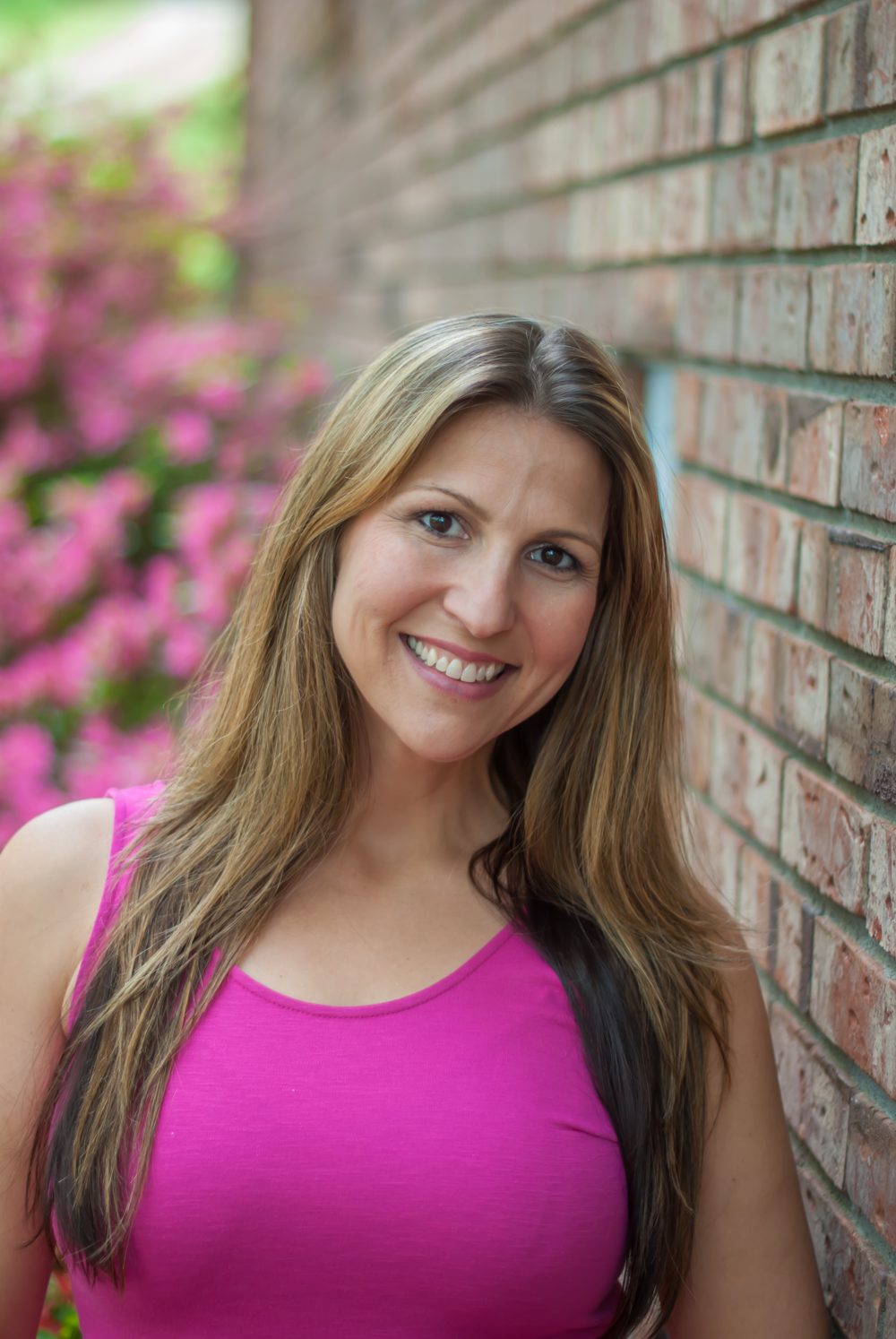 Serra Deville, founder of Georgia’s Dream Nannies, smiling outdoors by a brick wall with pink flowering bushes